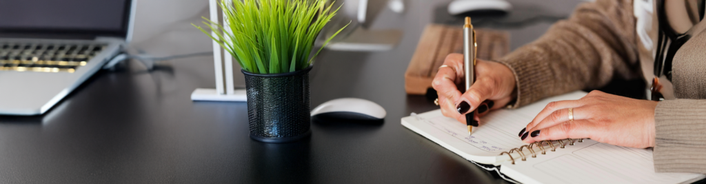 Close-up of a person writing in a notebook on a sleek black desk with a green potted plant, a mouse, and a laptop in the background.